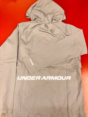 Grå vindjacka från Under Armour - Snygg grå vindjacka från Under Armour med huva och dragkedja framtill. Jackan har vita logotyper på bröstet och ryggen samt diskret #STORM-tryck på ärmen. Tillverkad i lätt, vattenavvisande material som passar perfekt för blåsiga dagar. Passar perfekt till sommaren. Helt ny oanvänd kommer med påse och tagg! Storleken är L men den är mindre i storlek passar mer som M.