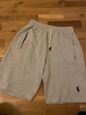 Randiga shorts från rlph - Säljer ett par snygga shorts från rlph med vit och grå smalrandig design. Shortsen har resårmidja med vit dragsko, två sidofickor med dragkedja och en bakficka med den klassiska häst-loggan broderad i svart. Perfekta för sommaren och riktigt sköna.