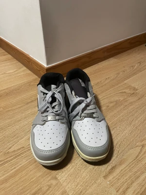 Nike Jordan 1 Low vit och grå - Nike Jordan 1 Low sneakers i vit och ljusgrå skinn med svart swoosh på sidan. Klassisk rund tå, perforerad tåbox och platt sula. Skorna har snörning och svart foder. Perfekt för dig som gillar stilrena och ikoniska sneakers.