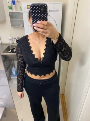 Svart spetsig croppad topp - Säljer en svart croppad topp i spets med långärmad design och djup v-ringning. Blusen har dekorativa kanter och transparenta ärmar i blommigt spetsmönster. Perfekt för dig som vill ha en edgy och trendig topp. 