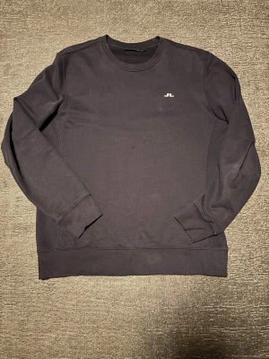 Svart sweatshirt från J.Lindeberg - Stilren svart sweatshirt från J.Lindeberg med rund halsringning och diskret logga på bröstet. Tröjan har långa ärmar, ribbade muddar och är tillverkad i mjukt bomullsmaterial. Perfekt för en avslappnad och trendig look.  Om du vill ha specifika bilder så är det bara att meddela!