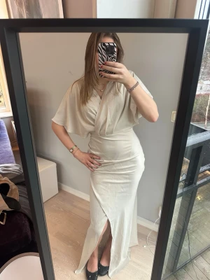 Ljus långklänning med omlott och slits - Säljer en elegant långklänning i ljus beige färg med omlott-design och v-ringning. Klänningen har korta, vida ärmar och en slits framtill som ger en snygg detalj. Perfekt för dig som vill ha en stilren och feminin look. Klänningen passar perfekt till en bal eller andra speciella tillfällen. Nypris är 1299!  Klänningen är ny och har prislapp, mitt pris- 900