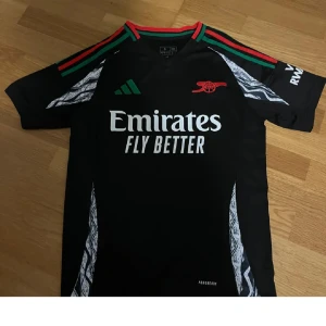Arsenal svart matchtröja Adidas - Snygg svart Arsenal matchtröja från Adidas med gröna och röda detaljer på axlarna, klubbmärke i rött och Emirates Fly Better-tryck på bröstet. Tröjan har korta ärmar och vita grafiska mönster längs sidorna. Perfekt för fotboll eller träning.