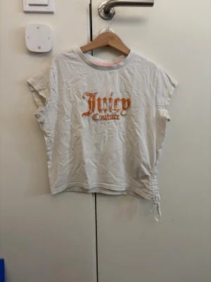 Vit Juicy Couture t-shirt med snörning - Vit t-shirt från Juicy Couture med orange logga framtill. T-shirten har korta ärmar och en unik snörningsdetalj på sidan. Tillverkad i mjuk bomull och har en avslappnad passform. Perfekt för en trendig och avslappnad stil.