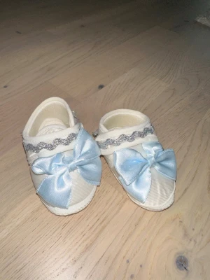 Söta babyskor med blå rosett - Supersöta babyskor i krämvitt tyg med stora ljusblå rosetter framtill och glittrigt silverband upptill. Perfekta för dop eller speciella tillfällen. Mjukt material och fina detaljer gör dem extra gulliga.