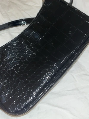 Svart croco-mönstrad axelväska - Snygg svart handväska med croco-mönstrad yta och glansig finish. Väskan har en avlång form och ett avtagbart axelband med silverfärgade metalldetaljer. Perfekt för dig som gillar trendiga accessoarer med lite edge.