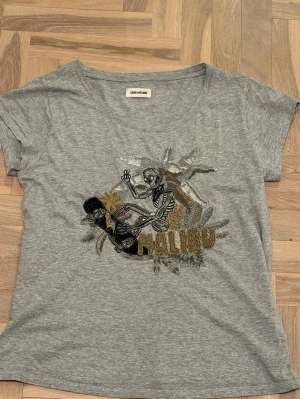 Grå t-shirt med paljettmotiv Malibu - Grå t-shirt från Zadig & Voltaire med coolt paljettmotiv av en skelettskatare och texten 'Malibu' i guld och silver. Kortärmad modell i mjuk bomull, perfekt för en avslappnad och trendig stil.