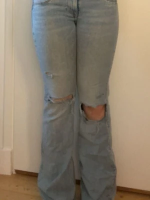 Ljusblå slitna bootcut jeans - Säljer ett par ljusblå bootcut jeans med slitningar och hål vid knäna. Jeansen har hög midja, klassiska fem fickor och är tillverkade i denim. Perfekta för en avslappnad och trendig look.