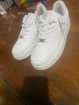 Nike Air Force 1 vita sneakers - Klassiska vita Nike Air Force 1 sneakers i lågt utförande. Skorna har en stilren design med perforerad tå, vita skosnören och metallfärgade detaljer vid snörningen. Tillverkade i läder med en tjock, vit sula och diskret Nike-logga på sidan.