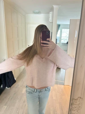 Ljusrosa stickad tröja med v-ringning - Mysig ljusrosa stickad tröja med bred v-ringning och långa, vida ärmar. Tröjan har en avslappnad passform och ribbstickade detaljer vid hals, ärmslut och nederkant. Perfekt att matcha med jeans för en chill look.