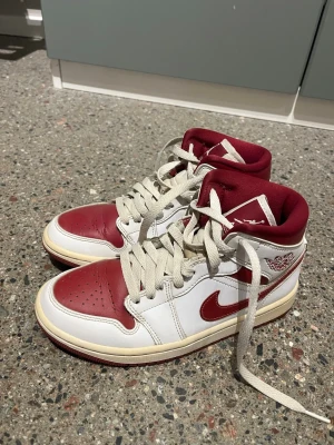 Nike Air Jordan 1 High röd/vit - Säljer ett par Nike Air Jordan 1 High sneakers i rött och vitt läder med klassisk vinge-logga på sidan och röd swoosh. Skorna har vita snören, röd insida och en retro-inspirerad sula. Perfekta för dig som gillar ikoniska basketskor och streetwear.
