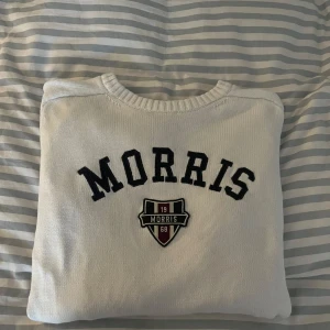 Vit stickad tröja från Morris - Snygg vit stickad tröja från Morris med svart broderad logga och emblem på bröstet. Tröjan har rund halsringning, ribbade muddar och lång ärm. Perfekt för dig som gillar klassisk och stilren look.
