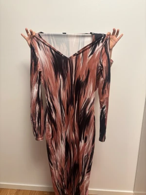 Mönstrad långklänning med v-ringning - Långklänning med abstrakt mönster i rosa, svart och beige toner. Klänningen har v-ringning och långa ärmar, tillverkad i ett mjukt och stretchigt material. Perfekt för dig som gillar unika prints och bekväma plagg.