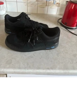 Svarta Nike Air Force 1 sneakers - Säljer ett par klassiska svarta Nike Air Force 1 sneakers med snörning och blå detaljer på hälen och sulan. Skorna har en låg profil och är tillverkade i skinn med Nike-logga på sidan och AIR-tryck på sulan.