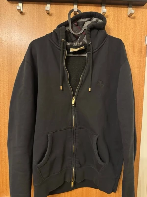 Svart hoodie från Burberry - Snygg svart hoodie från Burberry med dragkedja, huva och guldfärgade detaljer på dragkedjan och snörändarna. Tillverkad i mjuk bomull och har två stora fickor framtill. Perfekt för en chill och stilren look.