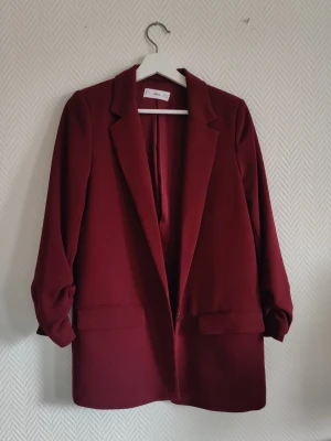 Vinröd kavaj från MNG - Stilren vinröd kavaj från MNG med klassisk krage. Kavajen har två fickor framtill och axelvaddar. Lite längre modell med 3/4 ärmar. Lite mer lila i tonen än bilderna visar.