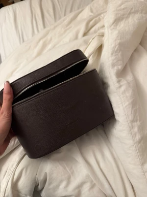 Mörkbrun necessär i läder - Snygg mörkbrun necessär från Caia. Väskan har ett praktiskt dragkedjelock och flera fack på insidan för smidig organisering av dina saker. Perfekt för att hålla ordning på småprylar eller smink i väskan.