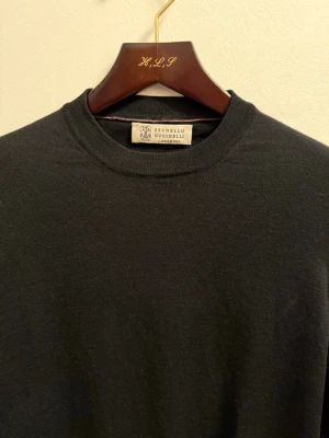 Brunello Cucinelli tröja  - Klassisk svart stickad tröja från Brunello Cucinelli i rundhalsad modell. Tröjan har långa ärmar, ribbade muddar och är tillverkad i ett mjukt, exklusivt material som ger en stilren och tidlös look.