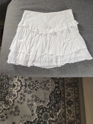 Vit volangkjol från Gina Tricot - Supersöt vit kortkjol från Gina Tricot med flera volanger och smock i midjan. Kjolen är luftig och har en flowy känsla, perfekt för varma dagar. Enkel att matcha med olika toppar för en fräsch look.