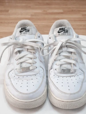 Nike Air Force 1 vita sneakers - Klassiska vita Nike Air Force 1 sneakers med lågt skaft. Skorna har en stilren design med perforerad tå, vita skosnören och Nike-logga på både plös och häl. Tillverkade i läder med gummisula för extra komfort och streetkänsla.