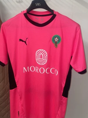 Marocko fotbollströja - Snygg rosa fotbollströja från Puma med Marockos landslagsemblem på bröstet och svart Puma-logga. Tröjan har svarta detaljer vid sidorna och halsen samt tryckt sponsorlogga på framsidan. Perfekt för dig som vill sticka ut på planen.