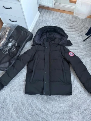 Svart dunjacka från Canada Goose - Säljer en svart dunjacka från Canada Goose med huva och klassisk logga på ärmen. Jackan har långa ärmar, dragkedja och knappar framtill samt fyra fickor. Perfekt för kalla dagar och riktigt snygg till vinterlooken.