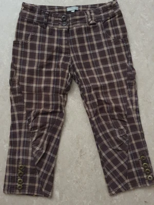 Byxor - Snygga bruna byxor med rutigt mönster i beige och mörkbrunt. Croppad modell med dekorativa knappar vid bensluten och dubbla knappar i midjan. Fickor både fram och bak. Perfekta för dig som gillar en unik och trendig stil.