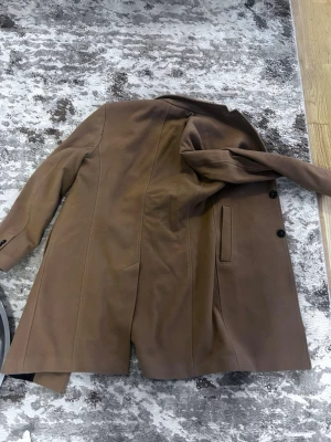 Brun kappa från Zara med bälte - Stilren brun kappa från Zara i storlek S. Kappan har ett avtagbart bälte i midjan, två sidofickor och knäppning med svarta knappar. Materialet är mjukt och ger en klassisk look, perfekt för kyligare dagar.