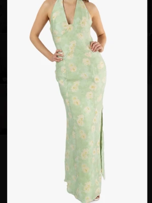 Grön blommig långklänning från adoore - Ljusgrön långklänning från adoore med blommigt mönster i vitt och gult. Klänningen har halterneck och en liten v-ringning framtill. Tillverkad i ett lätt och luftigt material, perfekt för varma dagar.