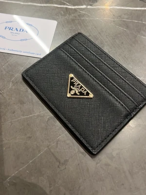 Svart kortfodral från Prada - Stilrent svart kortfodral från Prada i saffianoläder med ikonisk guldfärgad logga framtill. Fodralet har flera kortfack och en smidig, tunn design som passar perfekt i fickan. Lyxig och tidlös accessoar för dig som gillar exklusiva detaljer.