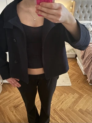 Kort mörk blå kappa med knappar - Säljer en stilren, kort kappa med knappar framtill och hög krage. Rocken har en boxig passform och raka ärmar, perfekt för dig som gillar minimalistisk och trendig stil. Passar till många olika outfits och är ett måste i garderoben. 