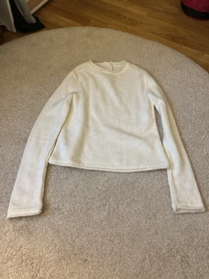 Vit långärmad tröja med knappar - Mysig vit långärmad tröja i mjukt material. Tröjan har rund halsringning och tre dekorativa knappar i nacken. Passar perfekt till lager på lager och har en enkel, stilren design.