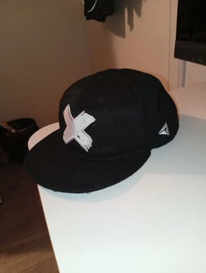 Svart keps från WLIKE med brodyr - Säljer en svart keps från WLIKE med en stor vit broderad X på framsidan och logga på sidan. Kepsen har platt skärm och justerbar baksida. Materialet är kraftigt tyg som ger en streetwear-vibe.