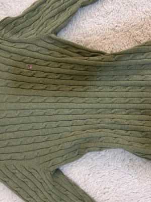 Olivgrön tröja GANT - Snygg olivgrön kabelstickad tröja från GANT i stretchig bomull. Tröjan har rund halsringning, långa ärmar och en liten broderad GANT-logga på bröstet. Passar dig som har storlek xxs-xs och S🤍