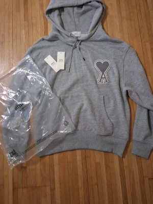 Grå hoodie från Ami Paris med hjärtdetalj (ÄKTA) - Snygg grå hoodie från Ami Paris med stor broderad hjärtdetalj och bokstaven A på bröstet. Hoodien har dragsko i huvan, ribbade muddar och känguruficka. Tillverkad i mjuk bomullsblandning för extra komfort. Perfekt för en avslappnad och trendig look. Pris kan diskuteras. Passar också M.