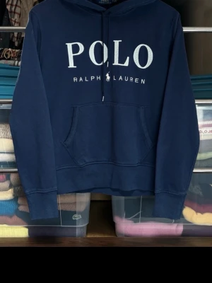Mörkblå hoodie från Polo Ralph Lauren - Mörkblå hoodie från Polo Ralph Lauren med stor vit logga på bröstet. Klassisk känguruficka framtill och justerbar huva med snören. Tillverkad i mjukt bomullsmaterial som känns skönt mot huden. Perfekt för en avslappnad och stilren look. Tröjan är helt ny, köptes för 1599 säljer för 650!