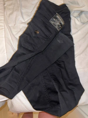 Svarta jeans från True Religion - Säljer ett par svarta jeans från True Religion med klassisk logga på bakfickan och snygga detaljer. Jeansen är bootcut och fickor med lock och knapp bak. Perfekta för dig som gillar street och premiumkänsla. Används ca 5 gånger och därav priset.