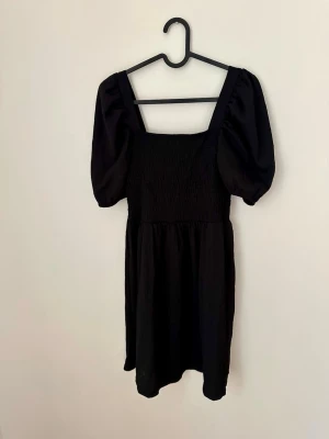 Svart kort klänning med puffärm -  svart kort klänning med puffärmar och fyrkantig ringning. Klänningen har smockad överdel 