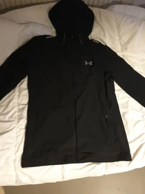Svart vindjacka från Under Armour - Svart vindjacka med huva från Under Armour. Jackan har dragkedja framtill, två sidofickor med dragkedja och en diskret logga på bröstet. Lätt och smidig modell som passar perfekt för blåsiga dagar. Materialet är tunt och funktionellt. Storlek S, passar bäst för 175-185, priset kan diskuteras vid snabb affär!