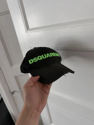 Svart Dsquared2 keps med neontext - Svart keps från Dsquared2 med böjd skärm och justerbart spänne bak. Framtill står det DSQUARED2 i neon-grön text och baktill DEAN & DAN CATEN. Kepsen är i bomull och har en cool, lite sliten look.