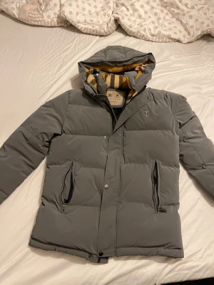 Grå pufferjacka från Burberry bara 1 hela världen  - Snygg grå pufferjacka från Burberry med klassiskt rutigt foder i gult, svart och vitt. Jackan har huva, dragkedja och tryckknappar framtill samt två fickor med dragkedja. Burberry-logga på bröstet och ärmen. Perfekt för kalla dagar.