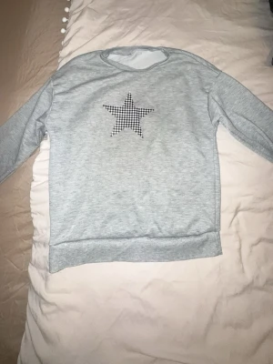 Grå sweatshirt med stjärna - Ljusgrå sweatshirt med rund hals och lång ärm. På bröstet finns en svartvit rutmönstrad stjärna som detalj. Perfekt för en avslappnad och trendig look.