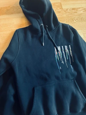 Svart hoodie från Amiri med tryck - Svart hoodie från Amiri med färgglatt droppande logotyptryck både fram och bak. Klassisk känguruficka och justerbar huva med snören. Mjuk och skön bomullskvalitet, perfekt för en avslappnad streetwear-look.