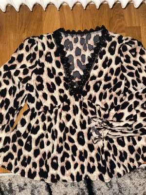 Leopardmönstrad blus med spets - Snygg leopardmönstrad blus med v-ringning och svart spetskant. Blusen har långa ärmar och ett knytband i midjan som ger en cool och feminin look. Materialet är lätt och mjukt, perfekt för att sticka ut i mängden.
