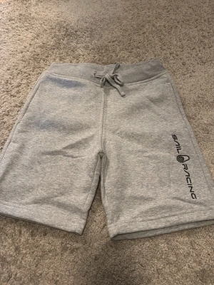 Grå shorts från Sail Racing - Snygga grå shorts från Sail Racing med logga längs ena benet. Mjukt material och bekväm passform med snörning i midjan. Perfekta för chill dagar eller sportiga aktiviteter.