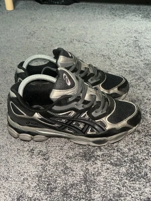 Svarta Asics sneakers med mesh och detaljer - Säljer ett par svarta Asics sneakers med silvriga och grå detaljer. Skorna har en chunky sula, mesh-ovandel för bra ventilation och klassiska Asics-loggan på sidan. Perfekta för dig som gillar sportig och trendig stil.