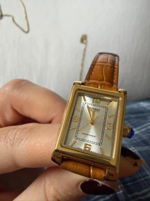Casio guld klocka med brun läderrem - Stilren Casio klocka med rektangulärt guldfärgat boett och ljus urtavla. Klockan har bruna läderremmar med krokodilpräglat mönster och guldfärgade detaljer. Perfekt accessoar för dig som gillar klassisk och tidlös stil.