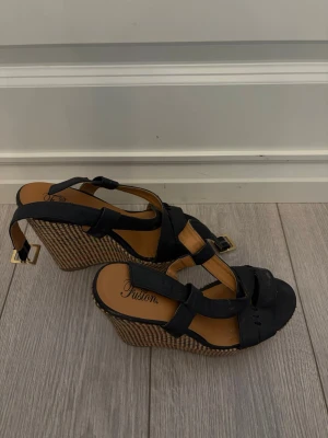 Svarta kilklackssandaler från Fusion - Snygga svarta sandaler från Fusion med kilklack i flätad bastlook. Remmar i svart konstläder och justerbart spänne i guld. Perfekta för dig som vill ha både höjd och komfort. Klassisk öppen tå och trendig design.