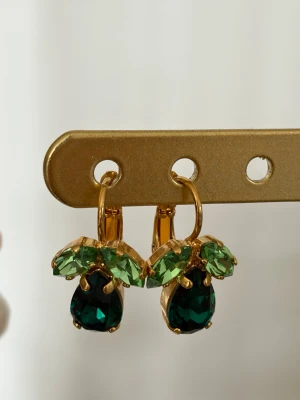 Guldörhängen med gröna stenar från Caroline svedbom - Snygga örhängen i guldfärgad metall med gnistrande gröna stenar i två nyanser. Stenarna är slipade och ger ett exklusivt intryck. Örhängena har en unik form som påminner om små blad eller bär och hängs i örat med en enkel krok.