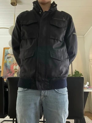 Woolrich jacka herr - Säljer en svart bomberjacka med fyra stora fickor framtill och ribbade muddar i midjan och vid ärmsluten. Jackan har hög krage och dragkedja framtill. Perfekt för dig som vill ha en stilren och funktionell jacka med streetkänsla.